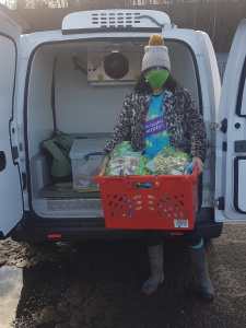 A volunteer in an Incredible Surplus t-shirt, face mask, and bobble hat unloads a crate of lettuces from the new electric refrigerated van.