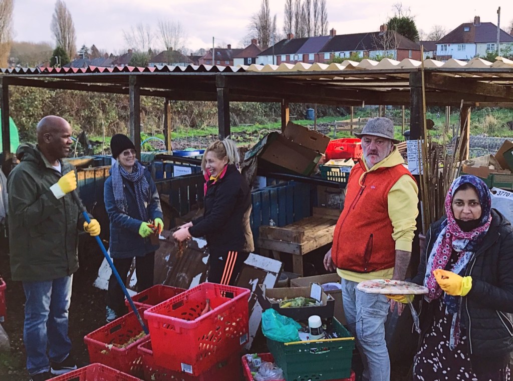 Five volunteers work on the allotment to sort the various types of waste for composting. Behind them is a covered area with bays separated by pallets.